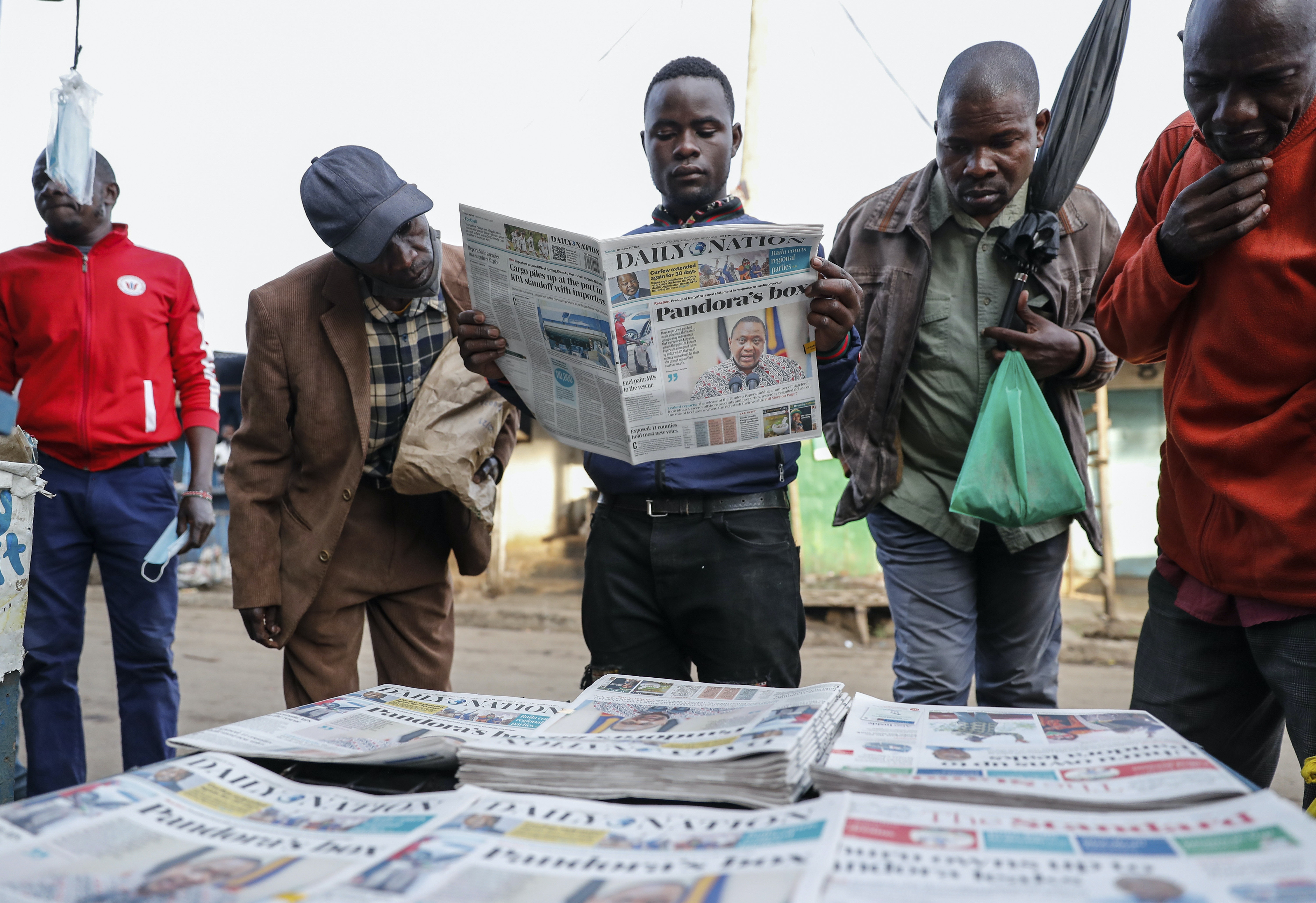 A group of men gather around a selection of newspapers, one of them reading one.